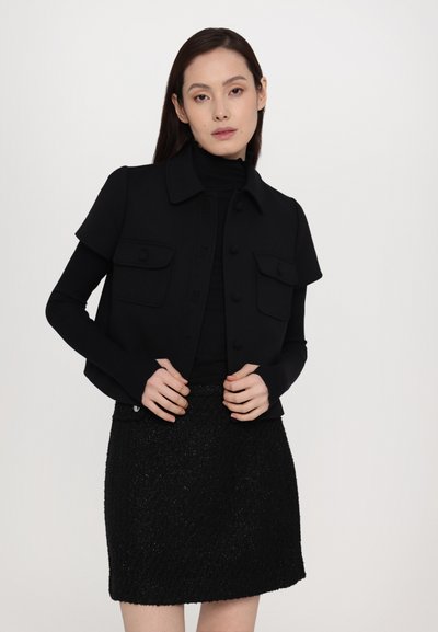 Black fitted jacket with short sleeves, front button closure, and two chest pockets, paired with a textured black mini skirt.