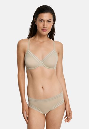 Donna che indossa reggiseno beige e biancheria intima abbinata con rifiniture in pizzo, in piedi contro uno sfondo chiaro uniforme.