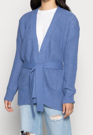 Femme portant un cardigan en maille bleu avec ceinture par-dessus une chemise blanche et un jean bleu clair déchiré sur un fond uni.