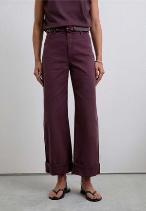 Pantalones de pierna ancha en algodón burdeos oscuro, con puños enrollados, presillas para el cinturón y un cinturón negro en contraste. Combinados con sandalias negras.