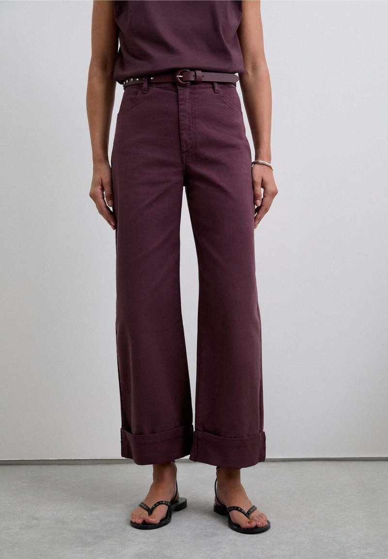 Pantalons évasés en coton bordeaux foncé, avec des ourlets roulés, des passants de ceinture et une ceinture noire contrastante. Portés avec des sandales noires.