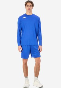 Långärmad blå sporttröja och matchande shorts, tillverkade av lättviktsmaterial. Har vit logotyp på bröstet och ovanför vänster knä.