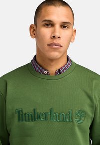 Felpa verde con logo "Timberland" ricamato e emblema dell'albero. Presenta un colletto a coste e un colletto di camicia a quadri sovrapposto.