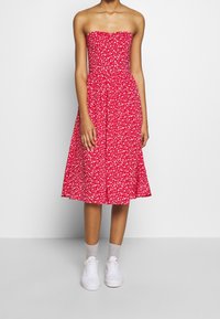 Punainen olkaimeton midi-mekko, jossa on valkoinen kukkakuosi, käytettynä vaaleanharmaiden sukkien ja valkoisten tennareiden kanssa seisovalla mallilla.