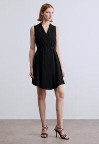 Μαύρο αμάνικο φόρεμα με V-λαιμόκοψη, εφαρμοστή μέση και φουσκωτή φούστα. Κατασκευασμένο από λείο ύφασμα· συνδυασμένο με μαύρα πέδιλα με λουράκια.