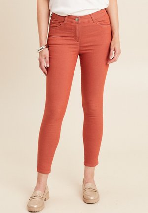 Jean skinny couleur rouille en coton extensible, avec cinq poches et une fermeture par bouton en métal. Texture lisse avec un design ajusté.