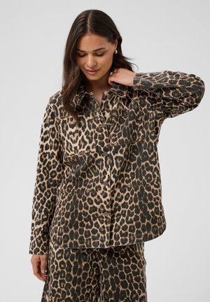 Vrouw draagt bijpassende jas en broek met luipaardprint, kijkt naar beneden met één hand die haar kraag raakt.