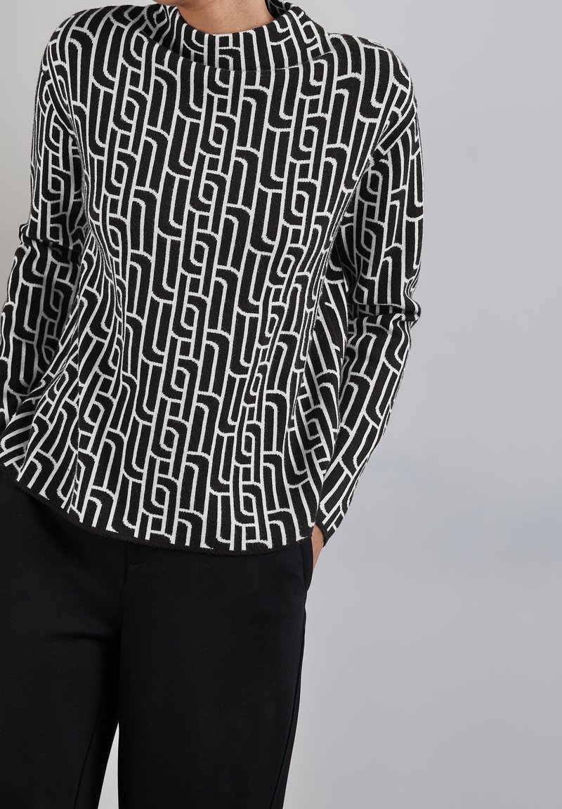 Pullover lavorato a maglia bianco e nero con un motivo geometrico, collo alto e maniche lunghe. La texture sembra morbida e leggermente strutturata.