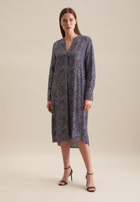 Robe à manches longues, marine avec un motif à pois blancs, décolleté en V et longueur mi-mollet. Associée à des sandales à talons hauts noires.