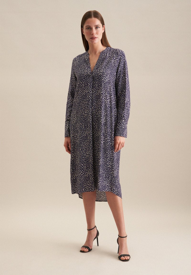 Robe à manches longues, marine avec un motif à pois blancs, décolleté en V et longueur mi-mollet. Associée à des sandales à talons hauts noires.