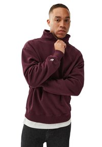 Burgundi ylisuurennettu collegepaitas, jossa on korkea kaulus, ribbaariset hihansuut ja helma. Vaatteen päällä on valkoinen paita, jossa on logo-etiketti hihassa.