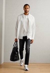 Cămașă cu dungi albe, peste un tricou alb simplu, blugi închiși la culoare și adidași albi. Poartă o geantă neagră. Fundal de podea din lemn.