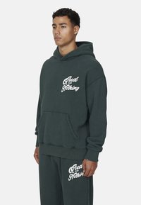 Felpa con cappuccio verde scuro in cotone, dotata di tasca frontale e testo bianco "Good for Nothing" sul petto e sulla gamba sinistra.