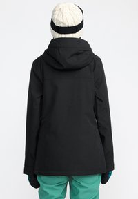 Schwarze wasserdichte Jacke mit Kapuze, die eine glatte Textur aufweist. Kombiniert mit hellblauen Hosen und einer kremfarbenen gestrickten Mütze. Die Handschuhe sind dunkel.