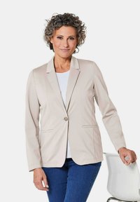 Beige op maat gemaakte blazer met geliseerde revers, enkele knoopsluiting en twee voorkezen, gecombineerd met een witte top en blauwe spijkerbroek.