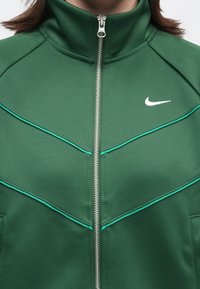Πράσινο αθλητικό μπουφάν με φερμουάρ και λευκό λογότυπο Nike, καθώς και ανοιχτό πράσινες λεπτομέρειες στην περιοχή του στήθους, φορεμένο από άτομο με καστανά μαλλιά.
