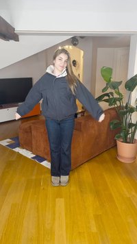 Sort puffjakke med lys grå hette, brukt over mørkeblå jeans. Står på tregulv i et oppholdsrom med planter og møbler.