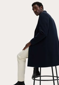 Marinblå ullrock med skräddarsydd passform, utrustad med notch-krage och sidofickor, i kombination med vita smala byxor och svarta ankelstövlar.