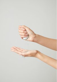 Selkeä geeli puristuu kädestä, valuen avoimeen kämmeneseen. Molemmat kädet ovat kevyesti kosteutettuja, tasaisella ihonsävyllä ilman näkyviä kuvioita.