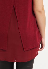 Vinröd topp med en delad bakdesign, lager på lager över ett genomskinligt tyg, med en texturerad stickad material och en avslappnad passform.