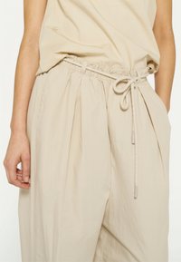 Pantaloni beige leggeri con vita elasticizzata e cordoncino. Presentano plissettature, tasche laterali e una texture liscia e morbida.