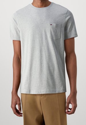 Mężczyzna w jasnoszarej koszulce z krótkim rękawem z kieszenią na piersi i małym logo, w połączeniu z jasnobrązowymi spodniami, na jednolitym tle.