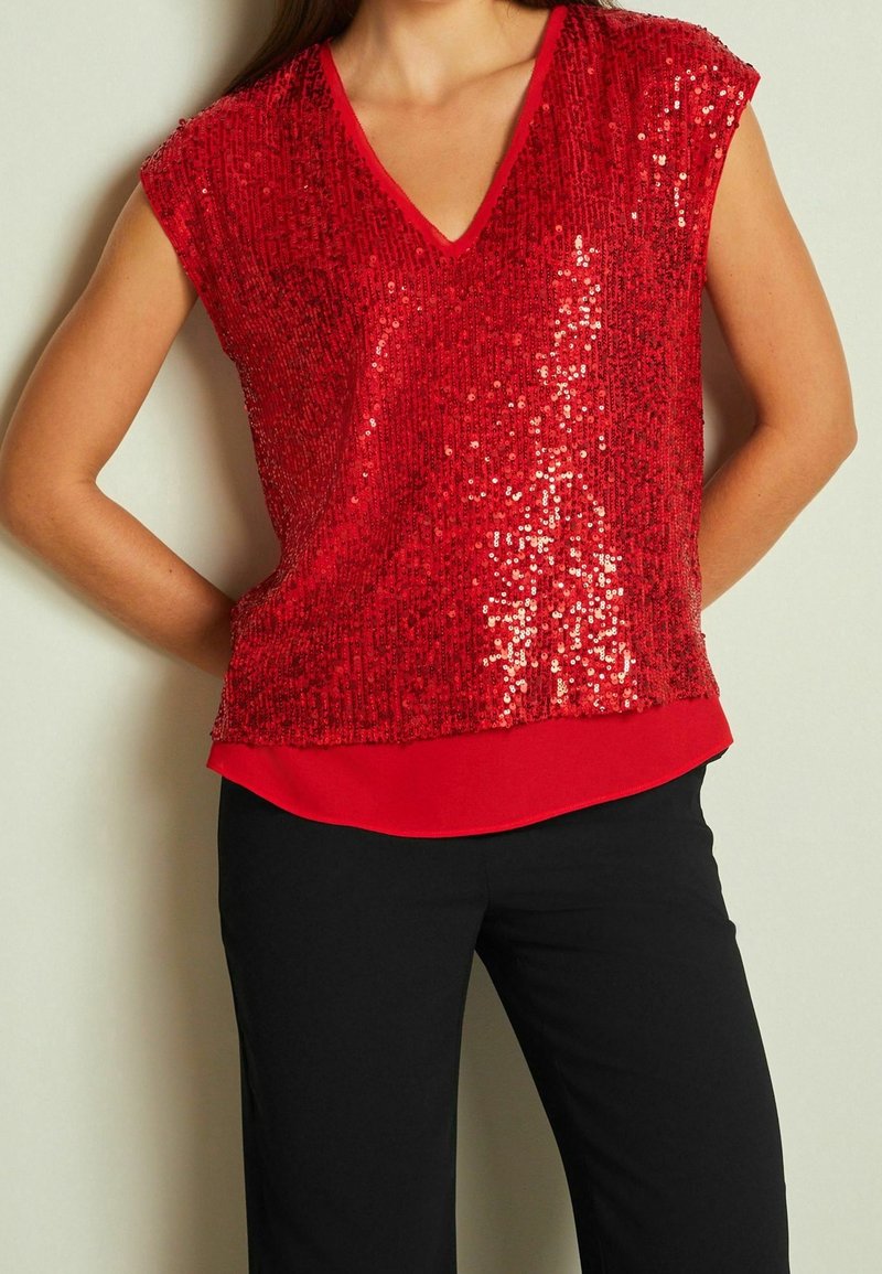 Femme portant un top sans manches rouge à sequins avec un col en V superposé à une chemise rouge, associé à un pantalon noir, debout les mains sur les hanches.
