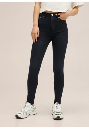 Person iført sorte højtaljede skinny jeans, hvid crop-top og hvide sneakers, stående med den ene hånd i lommen.