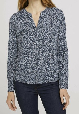 Frau trägt eine langärmlige Bluse mit blau-weißem abstraktem Muster, V-Ausschnitt und dunkelblaue Jeans.