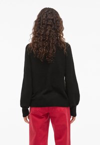 Hosszú, göndör barna hajú nő fekete pulóverben és piros nadrágban, háttal áll egy sima fehér háttér előtt.