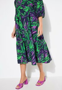Baumwollkleid mit dunkelblauem Grund, das auffällige grüne und lila Blätterdrucke aufweist. Kombiniert mit leuchtend lila Blockabsatzsandalen.