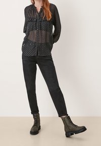 Blusa nera trasparente con pois, caratterizzata da una parte anteriore plissettata, abbinata a jeans neri slim-fit e stivali alla caviglia con lacci in verde oliva.