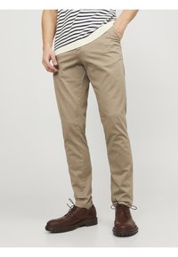 Uomo che indossa pantaloni beige slim-fit, scarpe stringate in pelle marrone e una camicia a righe, con una mano in tasca, in piedi su uno sfondo semplice.