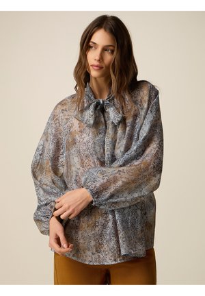 Kobieta o falowanych brązowych włosach, ubrana w prześwitującą, wzorzystą bluzkę z kołnierzykiem z kokardą i bufkami na rękawach, w połączeniu z musztardowymi spodniami.