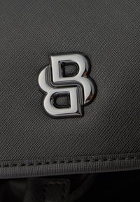 Pelēks teksturēts audums ar glancētu melnu logotipu, kurā ir savijumi ar burtiem "B" un "B", izšūtu apdari gar malām.