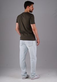 Mann in dunkelgrünem T-Shirt, hellblauen Jeans und weißenSneakern, der mit dem Rücken zu einem schlichten grauen Hintergrund steht.