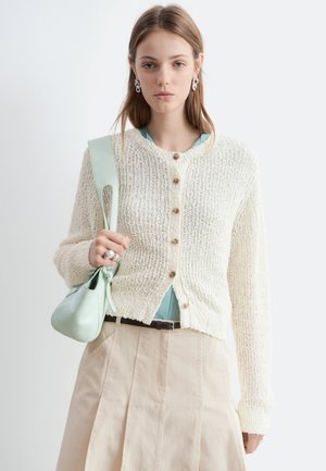 Jeune femme portant un cardigan en mailles crème sur un top bleu, jupe plissée beige, portant un sac à main vert clair sur l'épaule.