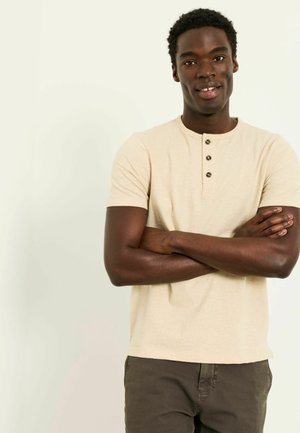 Young Black man standing with arms crossed, wearing a beige short-sleeve buttoned shirt and dark pants against a light background.