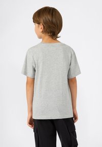 Jongen met lichtbruin haar, gekleed in een effen grijze korte mouwen T-shirt en zwarte cargobroek, met zijn rug naar de kijker op een witte achtergrond.