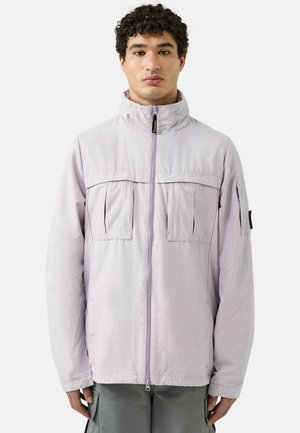 Hombre con chaqueta lavanda clara con cremallera, cuello alto y varios bolsillos, posando frente a un fondo blanco liso.