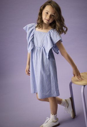 Young girl with curly hair wearing a blue striped dress and white sneakers, standing next to a wooden stool with lavender legs.