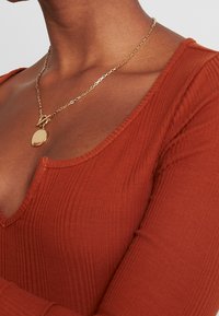 Collana in oro con pendente ovale, indossata sopra un top rosso ruggine testurizzato con scollatura a V profonda.