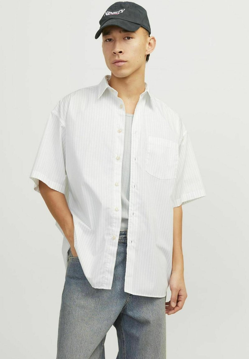 Chemise blanche à manches courtes avec des rayures verticales, poche poitrine unique, associée à un débardeur bleu clair et un jeans bleu délavé.