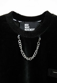 Suéter negro de canalé con cuello redondo, que presenta un detalle de cadena plateada y una etiqueta de marca en el interior. Tejido texturizado con un patrón vertical.