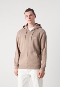 Felpa marrone con zip realizzata in tessuto morbido, dotata di una tasca frontale e di un piccolo logo ricamato sul lato sinistro in alto. Abbinata a pantaloni bianchi.