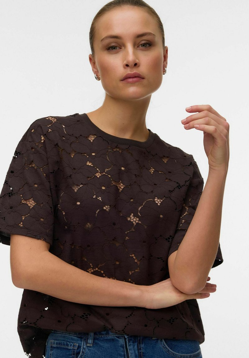 Femme à la peau claire et aux cheveux tirés en arrière porte un haut noir en dentelle florale à manches courtes et un jean bleu, posant avec un bras croisé et une main levée.