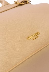 Borsa beige realizzata in materiale texturizzato, con logo e accessori in tonalità oro. Forma liscia e strutturata con dettagli di cucitura visibili.