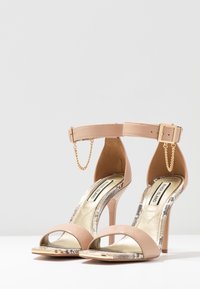 Beige högklackade sandaler med ankelrem, metalliska guldaccenter, lackat lädermaterial och en texturerad ormskinnsdetalj på sulan.