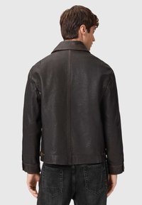 Giubbotto in pelle marrone con colletto classico, superficie testurizzata e dettagli con bottoni a scatto, abbinato a jeans scuri.