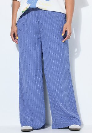 Person trägt lockere, hoch taillierte blaue Hosen mit vertikalen Streifen und Punkten, kombiniert mit weißen Schuhen und einem teilweise sichtbaren Grafik-T-Shirt.